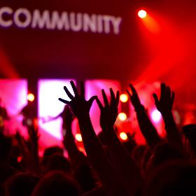 Alle feiern den Inhalts-Abschnitt (c) Photo by William White on Unsplash Man sieht die Hände einer feiernden Menschenmenge im rotem Licht. Das Wort 'Community' wird verschwommen an die Wand darüber projiziert.